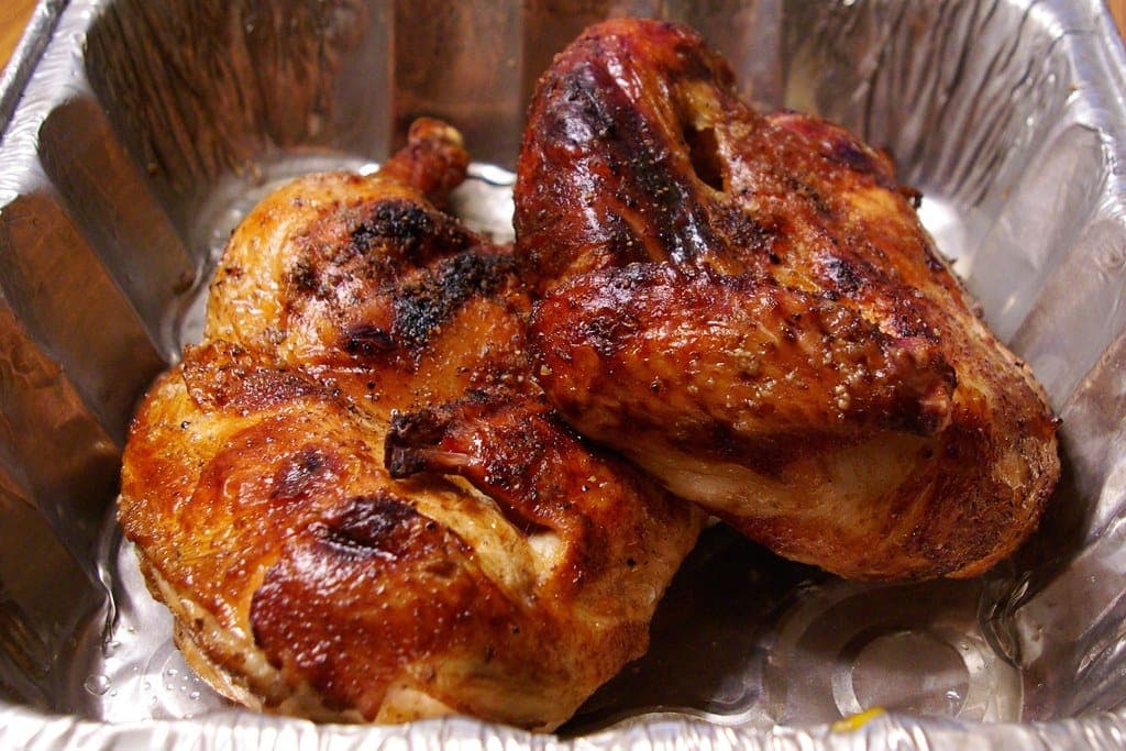 Two roast chicken halves with deep golden-brown, crisp skin in a foil tray