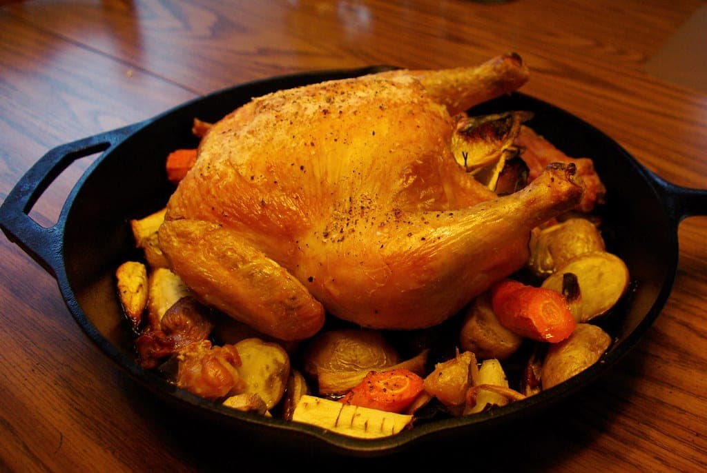 A golden roast chicken with roasted root vegetables in a cast iron skillet on a wooden table