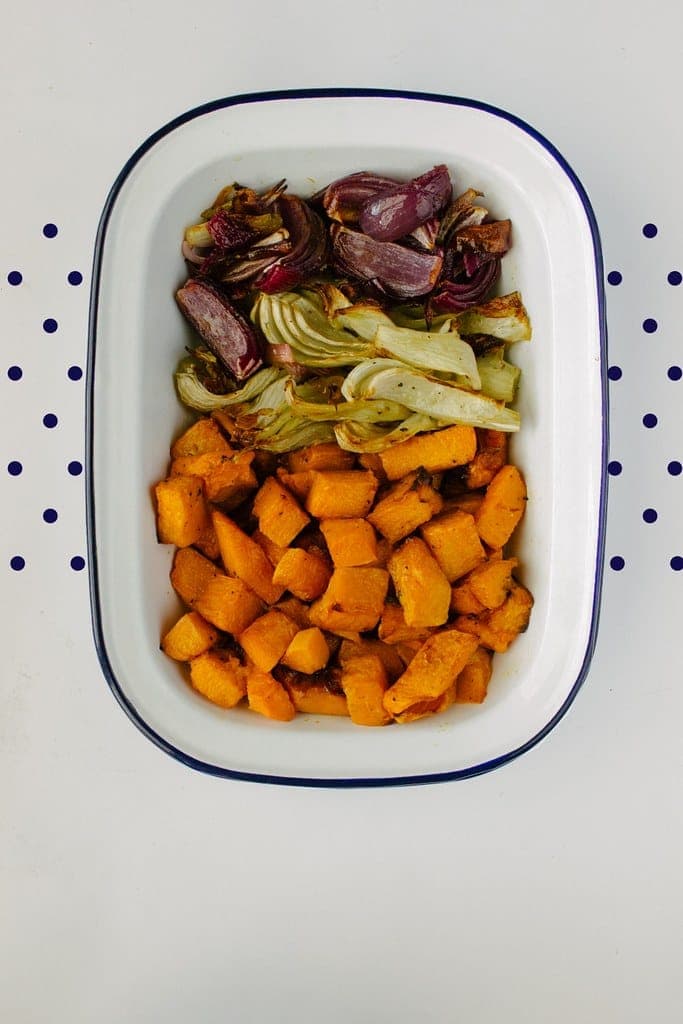 A white baking dish of golden roasted butternut squash cubes topped with roasted fennel and red onion