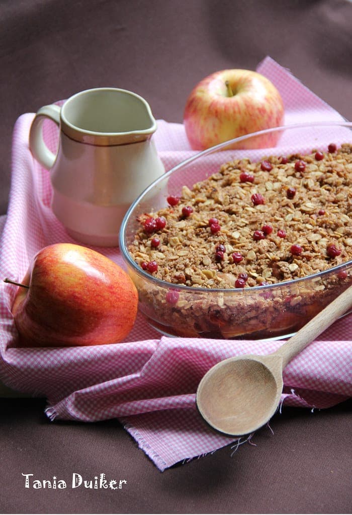 A glass baking dish of fruit crisp with a golden oat crumble topping dotted with red cranberries, next to a whole red apple