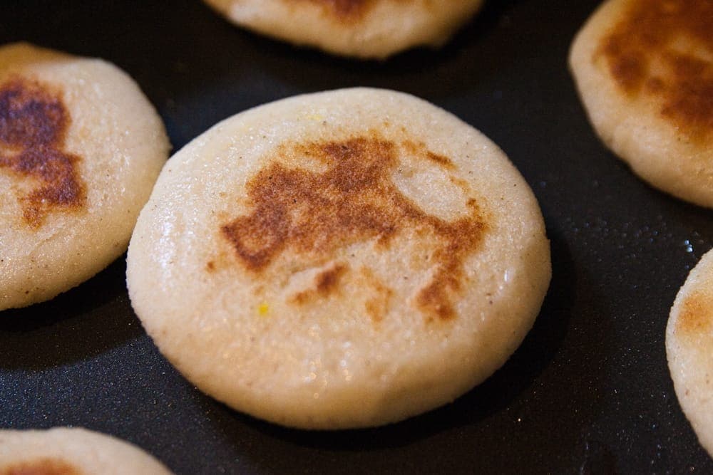 Round white corn arepas browning in a dark nonstick skillet