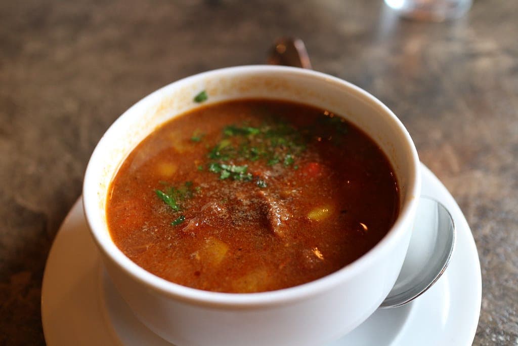 Cup of brothy red beef goulash with potato and chopped parsley on a saucer