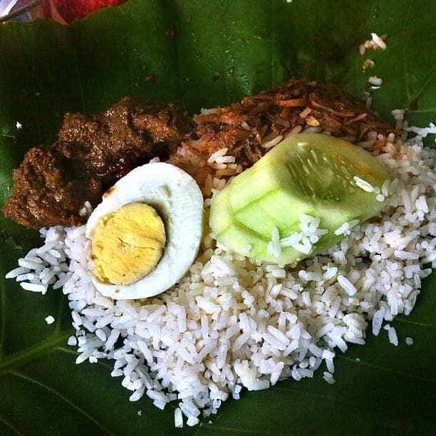 Dark beef rendang served with white rice, a halved boiled egg and cucumber on a banana leaf