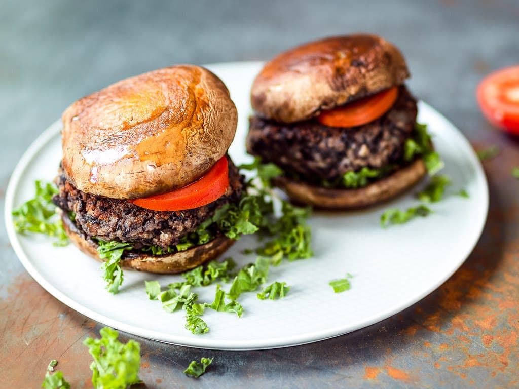 Two dark black bean burgers with tomato slices and shredded lettuce in glossy buns on a white plate