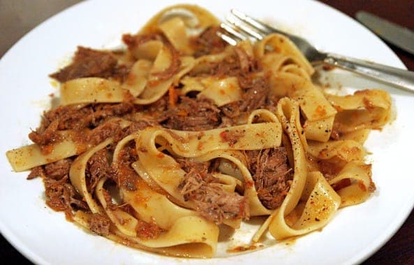 A plate of wide pappardelle ribbons tossed with shredded braised beef in a rich tomato sauce, with a fork on the rim
