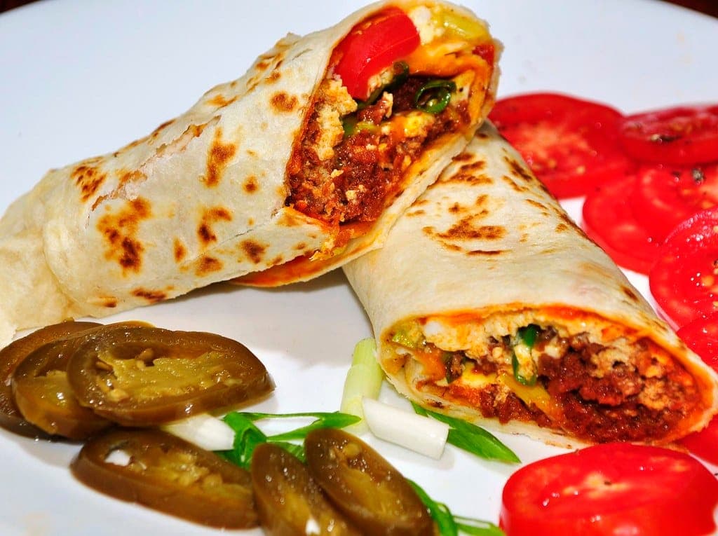 A breakfast burrito cut in half to show egg, chorizo and cheese, served with tomato and pickled jalapeño