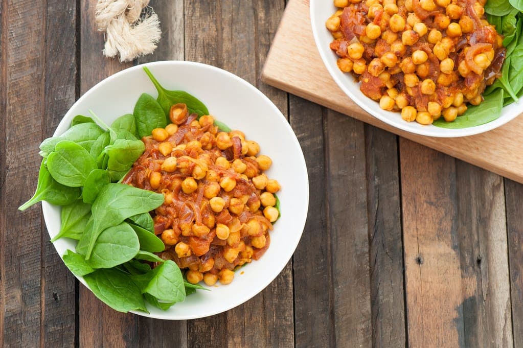 Bowls of spiced chickpea curry served with fresh baby spinach on a wooden table