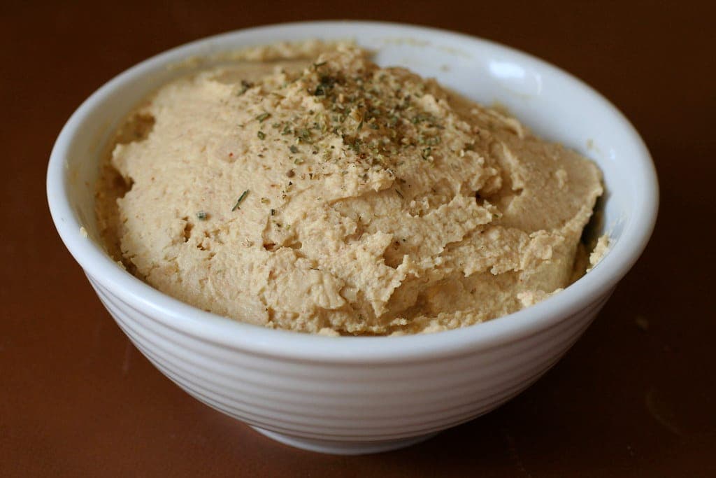 A white bowl filled with smooth, creamy hummus sprinkled with dried herbs