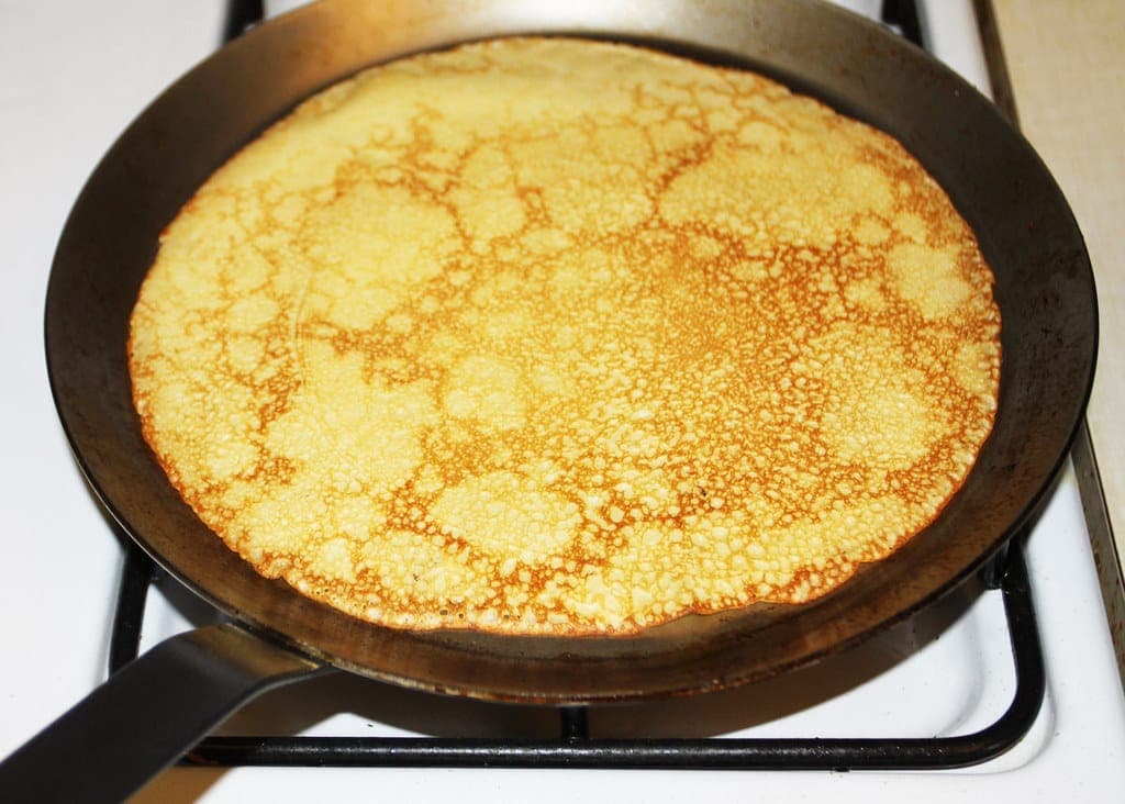 A thin golden crêpe cooking in a black crêpe pan on the stove