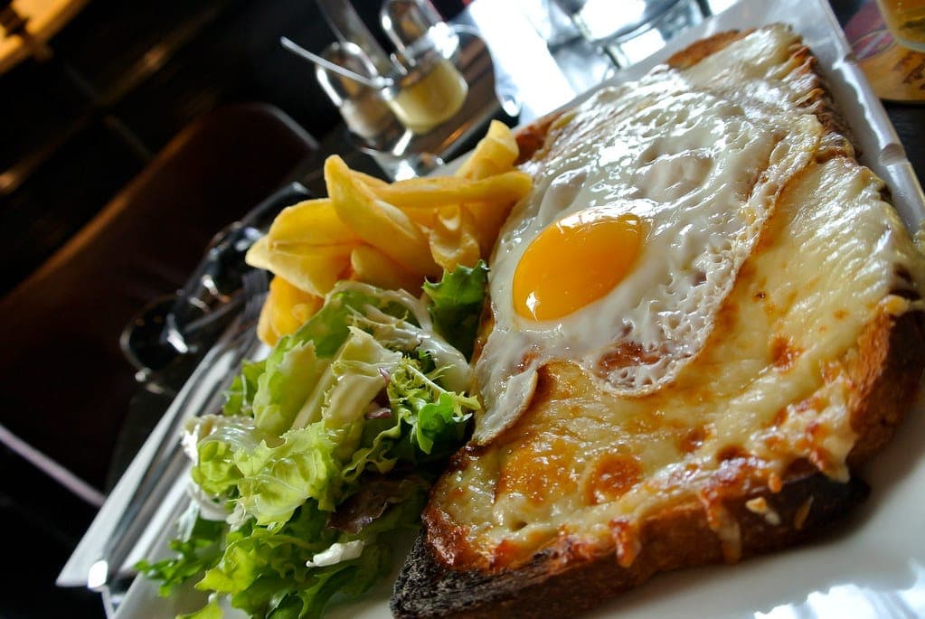 Croque madame with a browned, bubbling cheese top and a fried egg, served with salad and fries