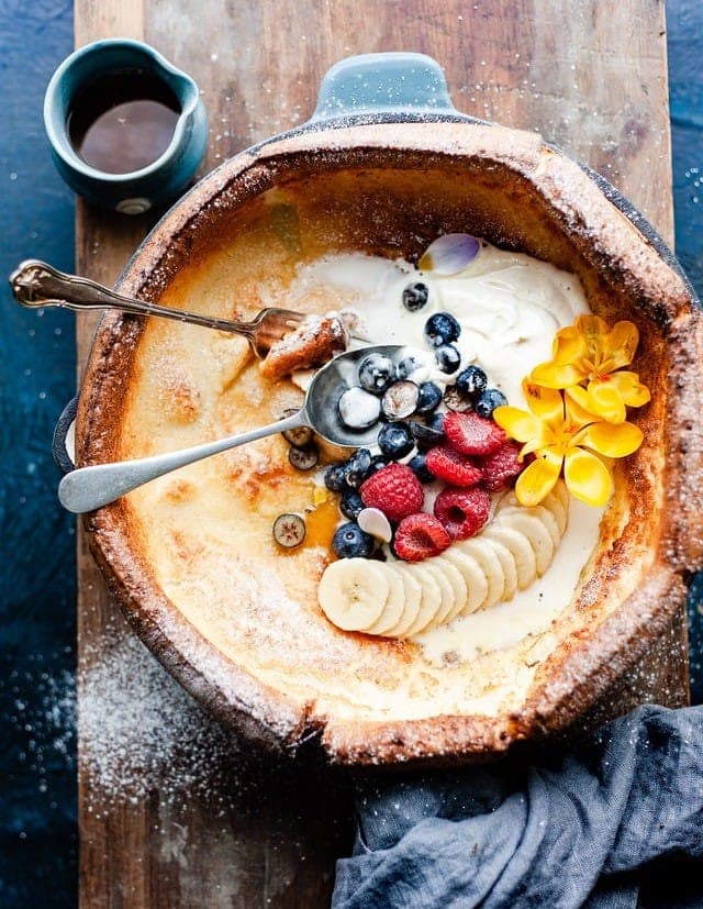 A puffed Dutch baby pancake in a skillet topped with yogurt, berries, banana and yellow flowers