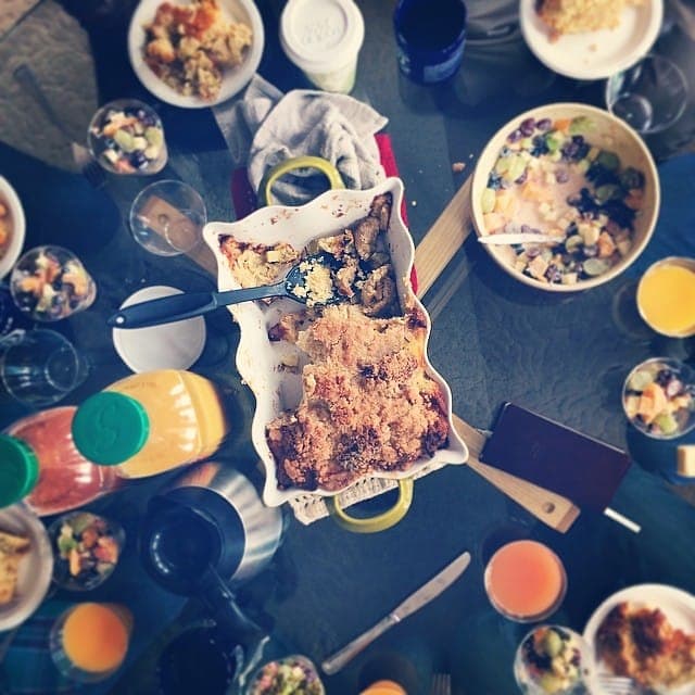 A crumb-topped breakfast bake in a white baking dish on a brunch table with fruit salad and juice