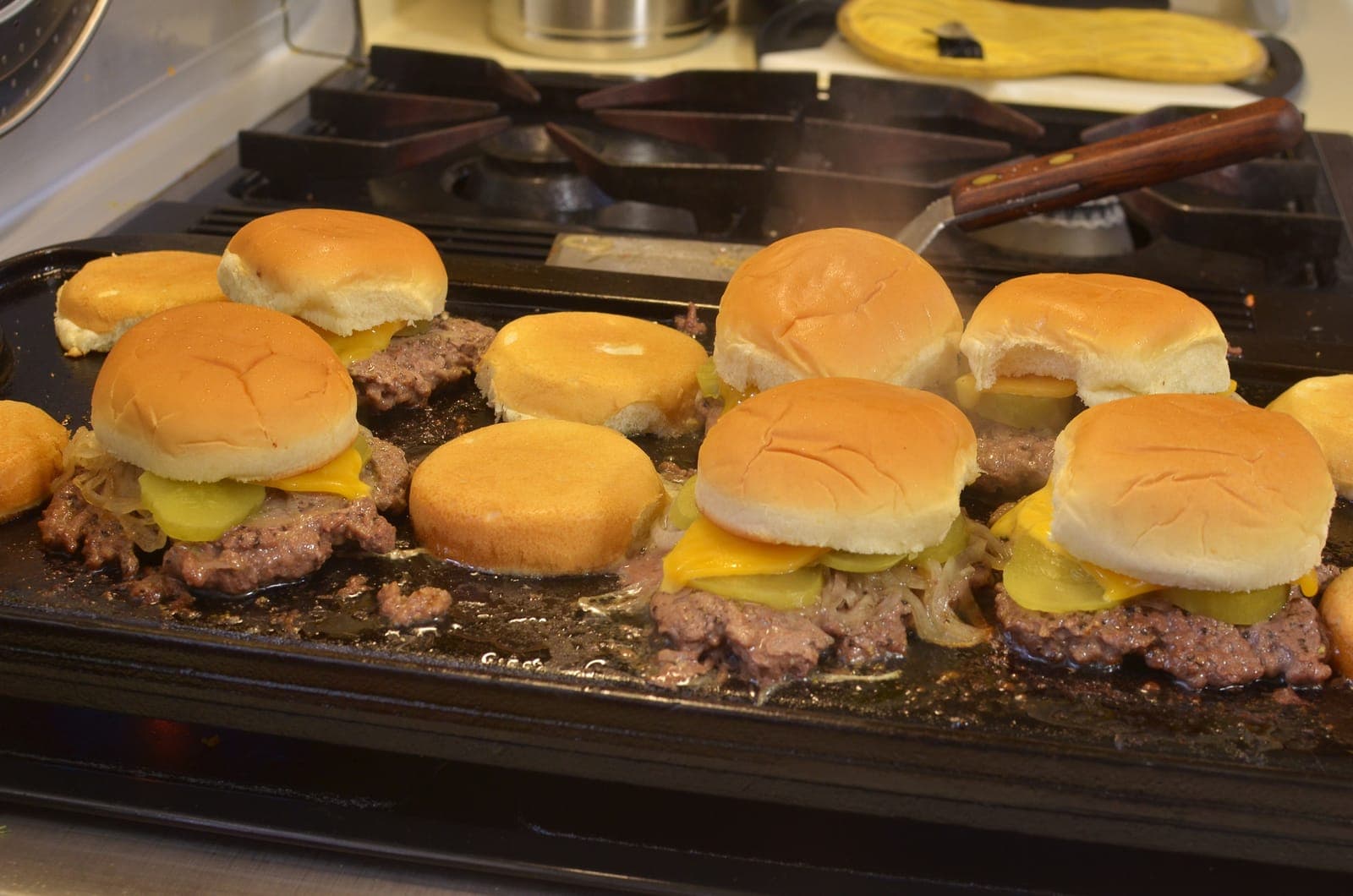 A batch of cheese-topped sliders with soft rolls cooking together on a griddle