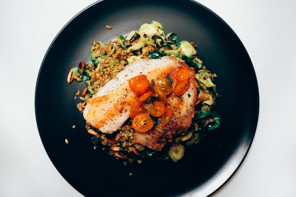 Pan-seared white fish fillet with kumquats over a herby grain and vegetable mix on a black plate