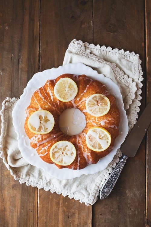 A golden Bundt cake drizzled with white glaze and topped with thin lemon slices, on a white plate over a linen cloth