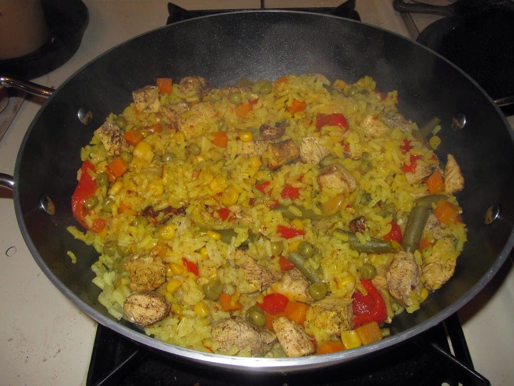 Large skillet of saffron-yellow rice with chicken pieces, peas, peppers and corn