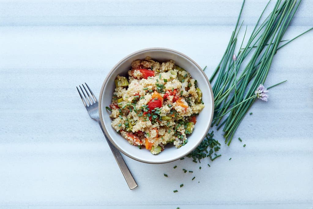 Bowl of quinoa salad with chopped vegetables beside a fork and fresh chives