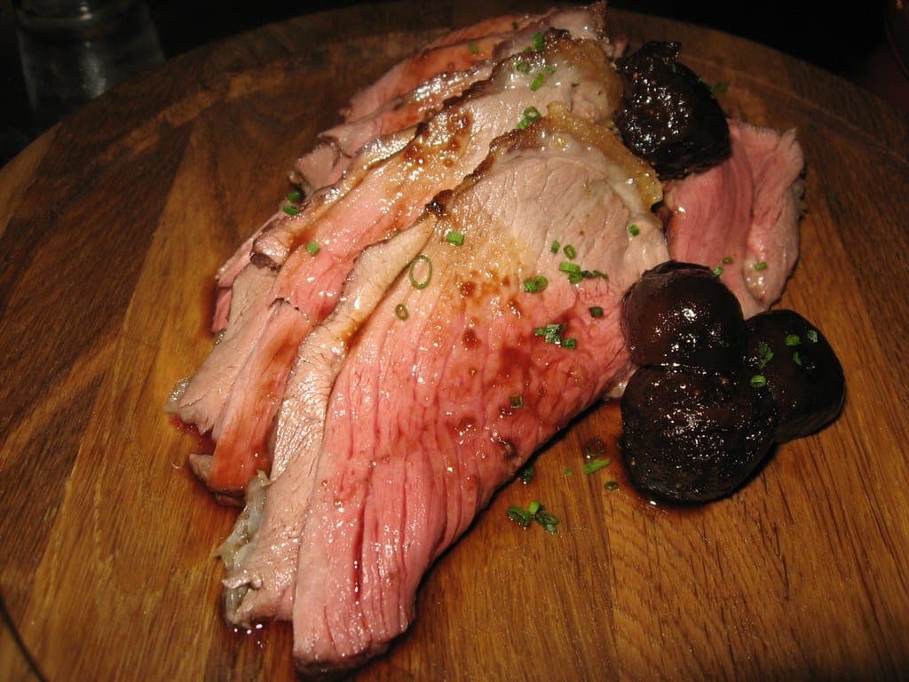 Slices of pink roast lamb on a wooden board with roasted shallots