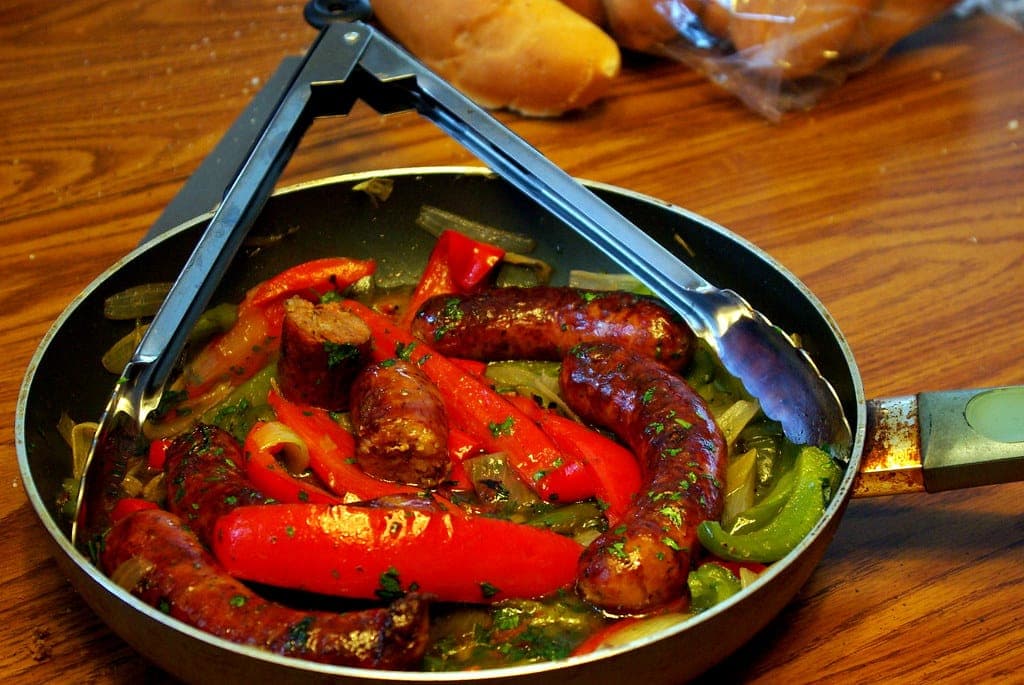 Skillet of browned sausages with red and green peppers and onions, with tongs
