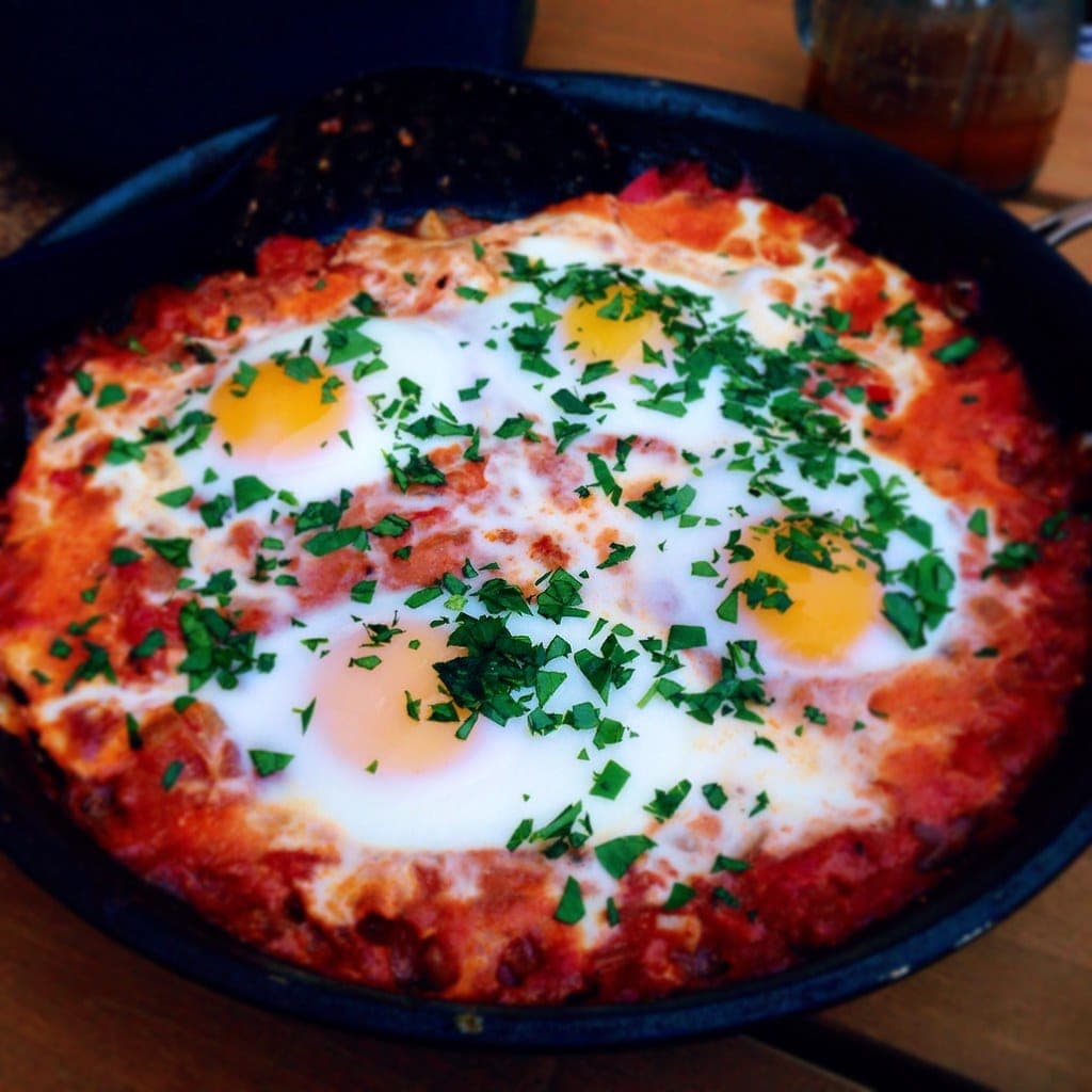 A cast-iron skillet of shakshuka with several eggs nestled in a red tomato and pepper sauce, sprinkled with chopped parsley