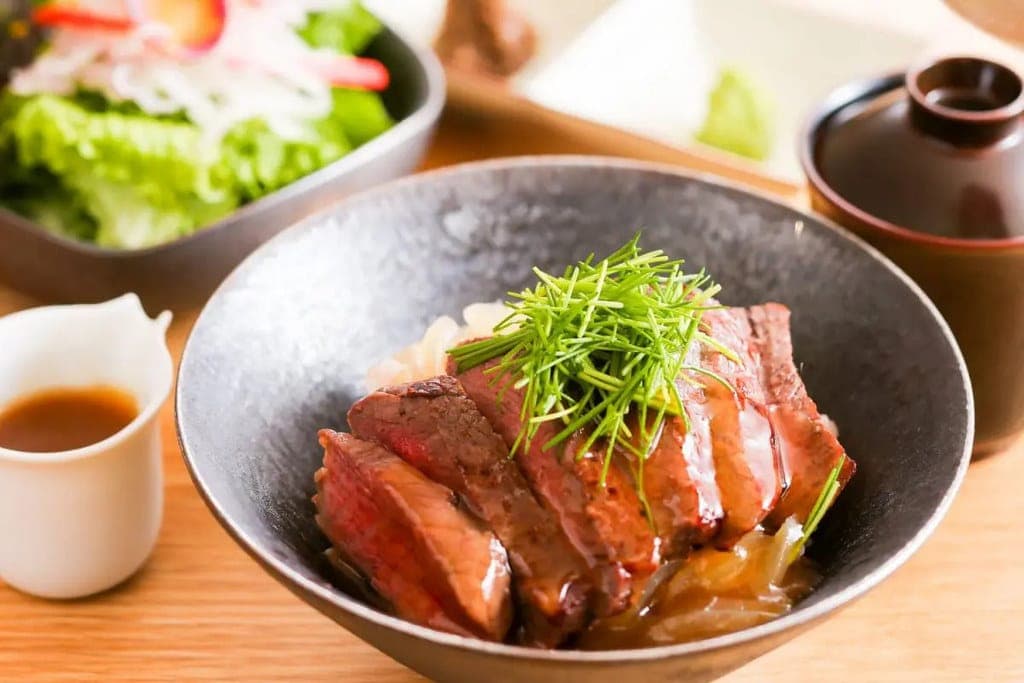 Sliced seared steak over rice in a dark bowl, topped with fine scallion strands