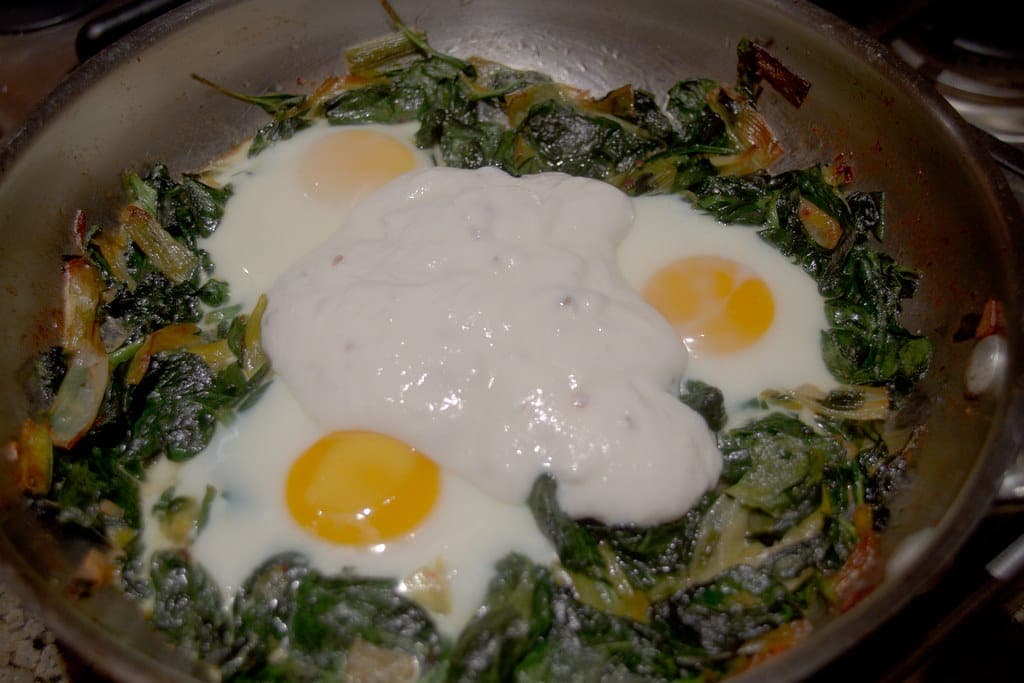 Eggs cooked over wilted spinach and topped with spoonfuls of yogurt in a pan