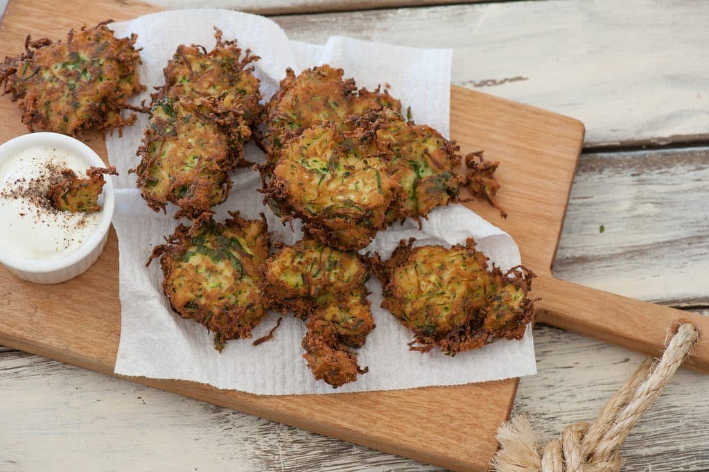 Crispy zucchini fritters on paper towel on a wooden board with a bowl of yogurt sauce